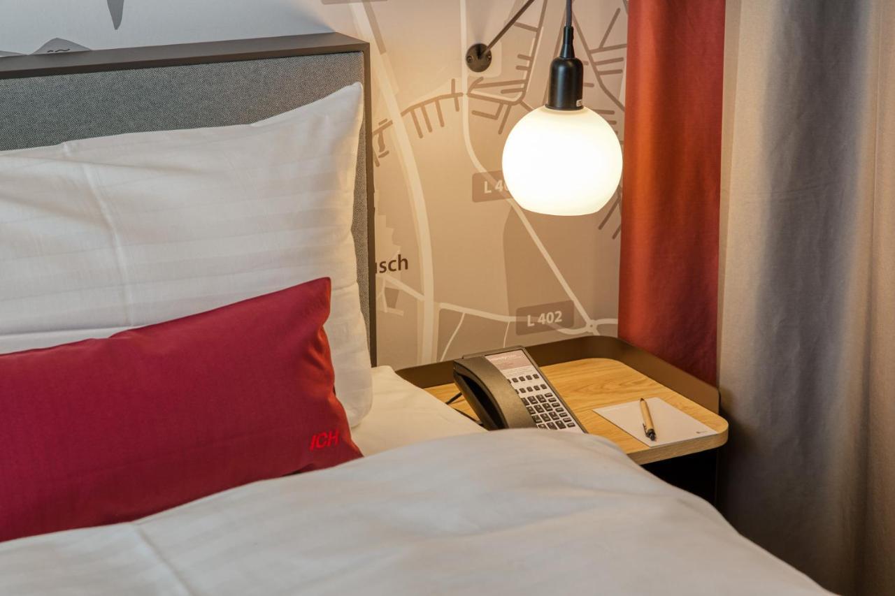 Intercityhotel Berlin Airport Ber Terminal 1 & 2 Schoenefeld Ngoại thất bức ảnh The photo shows a cozy bedroom setup. There's a neatly made bed with white bedding and a red decorative pillow. Beside the bed, a small bedside table holds a telephone and a notebook or notepad, along with a pen. A round pendant light hangs above the